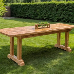 以下が含まれることがあります： 芝生の上に置かれた、自然な仕上がりの大きな長方形の木製テーブル。 テーブルには頑丈な脚と、緑の植物が入った装飾的なプランターボックスがあります。 テーブルの長さは約244cmです。