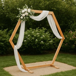 Op de afbeelding: Houten zeshoekige trouwboog versierd met witte stof en bloemen. De boog staat buiten op een grindoppervlak, met groen op de achtergrond. De boog is gemaakt van lichtgekleurd hout.