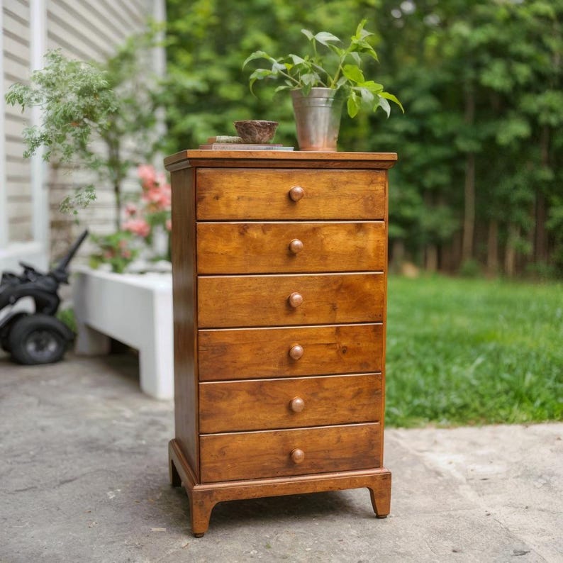 May include: A tall, wooden chest of drawers with six drawers and round wooden knobs. The chest is a warm, medium brown color and sits on short legs. A small metal bucket with a plant and a small wooden bowl are on top.