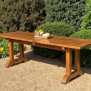 Może przedstawiać: Duży, prostokątny drewniany stół o ciepłym, czerwonobrązowym wykończeniu. Stół ma podstawę w kształcie kozła i stoi na zewnątrz na żwirowej powierzchni, otoczony zielenią. Na stole znajduje się mała taca z jedzeniem.