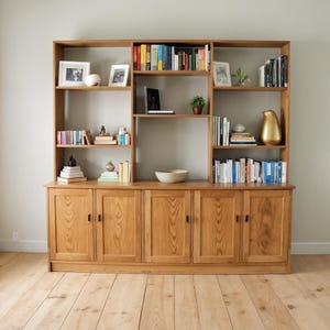 Puede incluir: Una gran librería de madera clara con múltiples estantes y armarios. La librería está llena de libros, objetos decorativos y fotos enmarcadas. La madera tiene un acabado natural y el diseño es clásico y funcional.