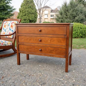 Op de afbeelding: Een houten borst met drie laden en een warme bruine afwerking. Elke lade heeft een ronde, donkerbruine knop. De borst staat op vier poten en staat buiten in de buurt van een schommelstoel met bloemenkussens.