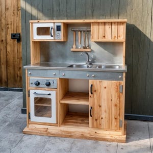 Puede incluir: Una cocina de juguete de madera con microondas, horno, fregadero y almacenamiento. La cocina tiene una encimera de acero inoxidable y un protector contra salpicaduras de madera. La puerta del horno y la del microondas son blancas.