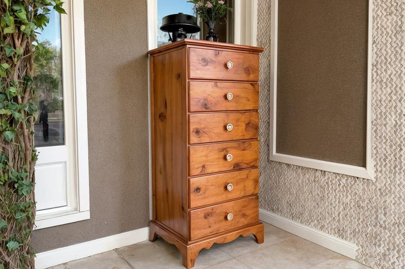 May include: A tall, wooden chest of drawers with six drawers. The chest is made of light-colored wood with a warm, reddish-brown tone. Each drawer has a round, light-colored knob. The chest is standing on a stone surface.