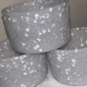 May include: A stack of three frosted gray candle holders. Each holder is decorated with white snowflake designs and glitter. The holders are cylindrical with a slightly flared top. The overall theme is winter or holiday decor.