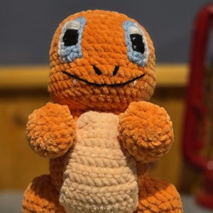 May include: A handmade orange crochet plush toy with a light peach belly. The toy has black eyes with blue accents, a smiling mouth, and white details on its feet. The plush is standing on a silver base.