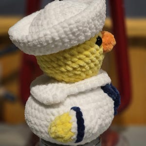 May include: A plush crochet duckling wearing a white sailor hat and a white sailor collar with dark blue trim. The duckling has a yellow head and body, a small orange beak, and black eyes. The texture is soft and fuzzy, crafted from yarn.