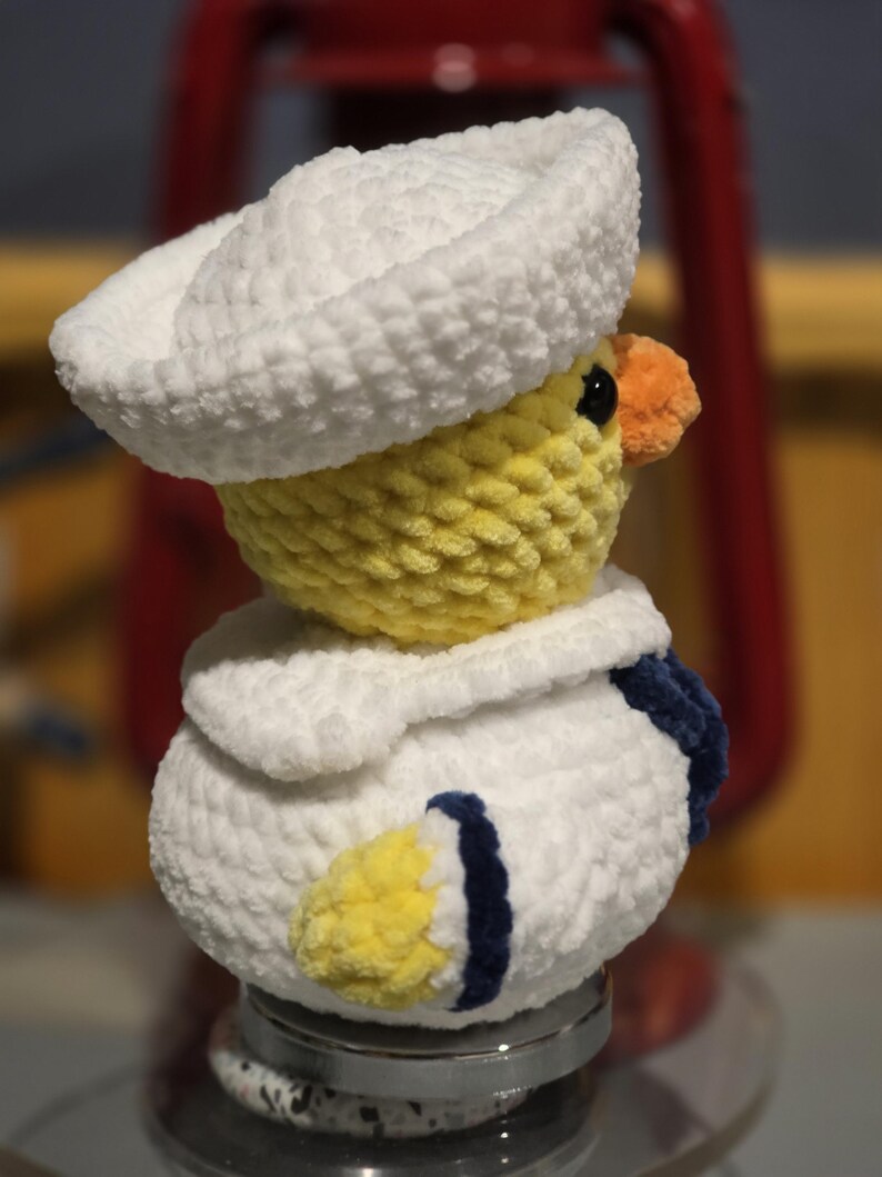 May include: A plush crochet duckling wearing a white sailor hat and a white sailor collar with dark blue trim. The duckling has a yellow head and body, a small orange beak, and black eyes. The texture is soft and fuzzy, crafted from yarn.