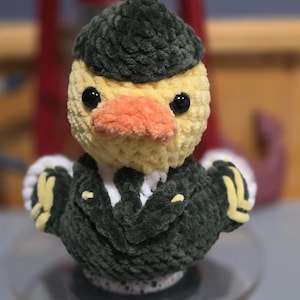 May include: A crocheted duck plushie wearing a dark green military-style uniform with yellow accents and a matching dark green cap. The duck has a pale yellow head, a soft orange beak, and black plastic eyes, crafted from textured yarn.