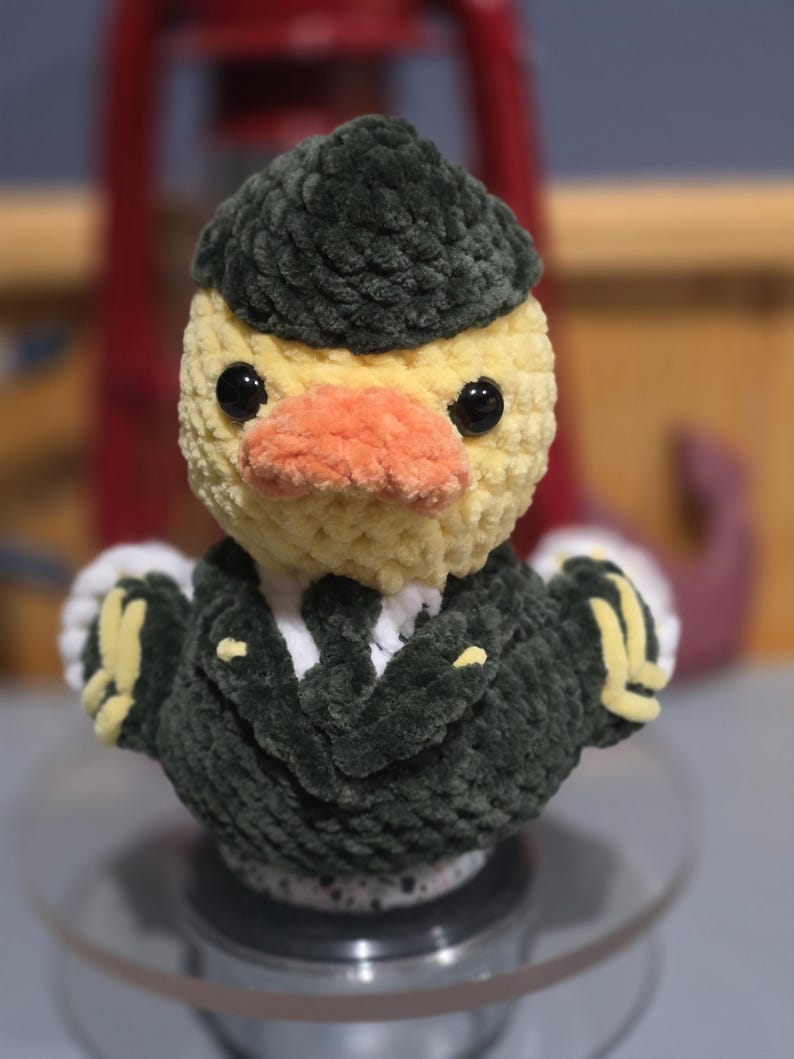 May include: A crocheted duck plushie wearing a dark green military-style uniform with yellow accents and a matching dark green cap. The duck has a pale yellow head, a soft orange beak, and black plastic eyes, crafted from textured yarn.