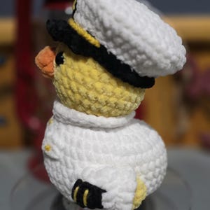 May include: A crocheted plush duckling wearing a white captain's hat with a black brim and a white uniform jacket. The duckling has yellow yarn, a small orange beak, and black details on the hat and sleeves. The texture is soft and fuzzy.