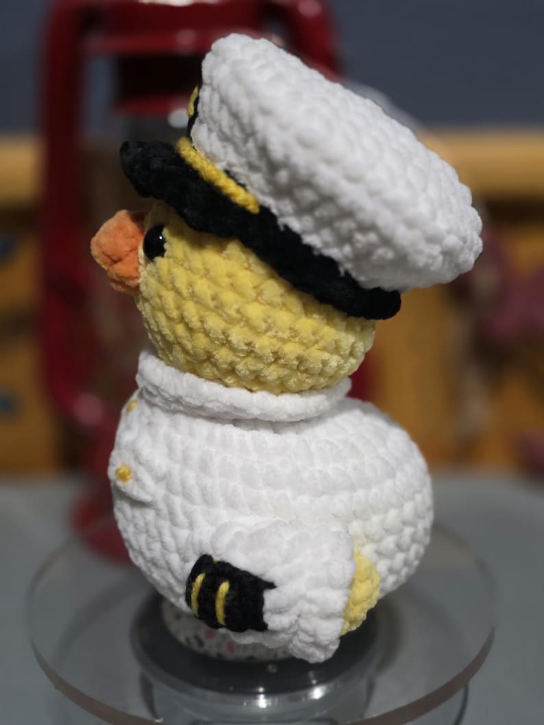 May include: A crocheted plush duckling wearing a white captain's hat with a black brim and a white uniform jacket. The duckling has yellow yarn, a small orange beak, and black details on the hat and sleeves. The texture is soft and fuzzy.