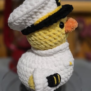 May include: A crocheted plush duck wearing a white captain's hat with a black brim and yellow trim. The duck has a yellow body, an orange beak, and a white uniform shirt featuring black cuffs with yellow stripes on the sleeves.