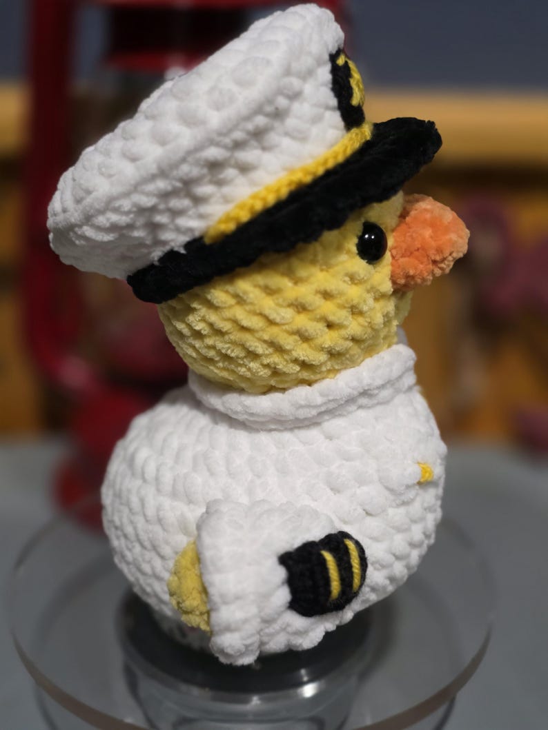May include: A crocheted plush duck wearing a white captain's hat with a black brim and yellow trim. The duck has a yellow body, an orange beak, and a white uniform shirt featuring black cuffs with yellow stripes on the sleeves.