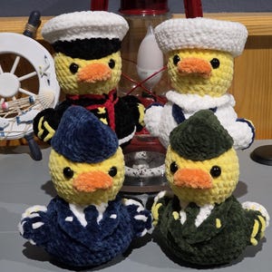 May include: Four crocheted plush ducks dressed in various military-style uniforms. Two ducks wear white sailor hats, while the others wear dark blue and green garrison caps. Each duck has a yellow body, orange beak, and black plastic eyes.