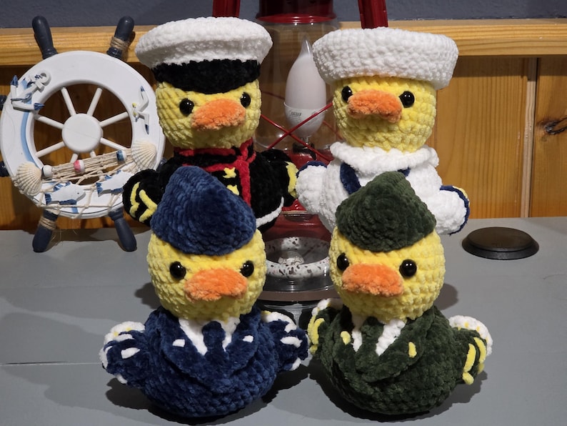 May include: Four crocheted plush ducks dressed in various military-style uniforms. Two ducks wear white sailor hats, while the others wear dark blue and green garrison caps. Each duck has a yellow body, orange beak, and black plastic eyes.