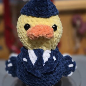 May include: A crocheted plush duck wearing a dark blue pilot hat and a matching dark blue uniform jacket with a white shirt and tie underneath. The duck has a yellow head, a textured orange beak, and two round black eyes.