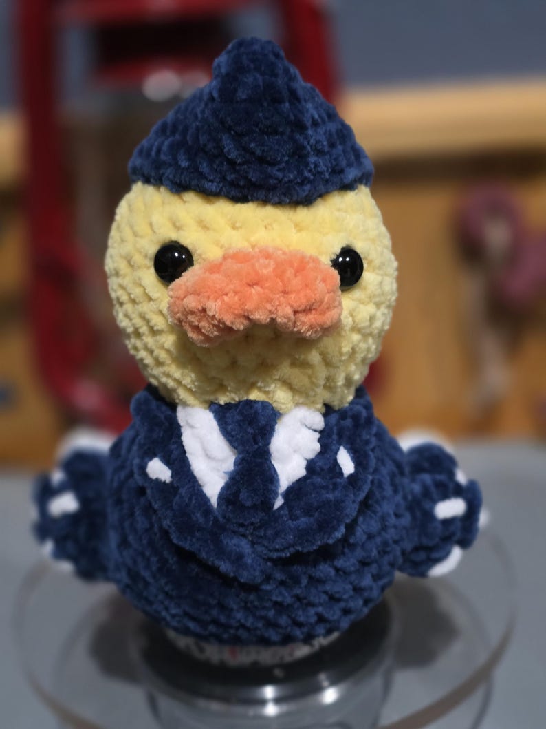 May include: A crocheted plush duck wearing a dark blue pilot hat and a matching dark blue uniform jacket with a white shirt and tie underneath. The duck has a yellow head, a textured orange beak, and two round black eyes.