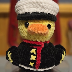 May include: A crocheted plush duck wearing a white and black sailor hat, a black jacket with yellow accents, a red scarf, and an orange beak. The duck has black plastic eyes and is crafted from soft, textured yarn in a standing, upright position.