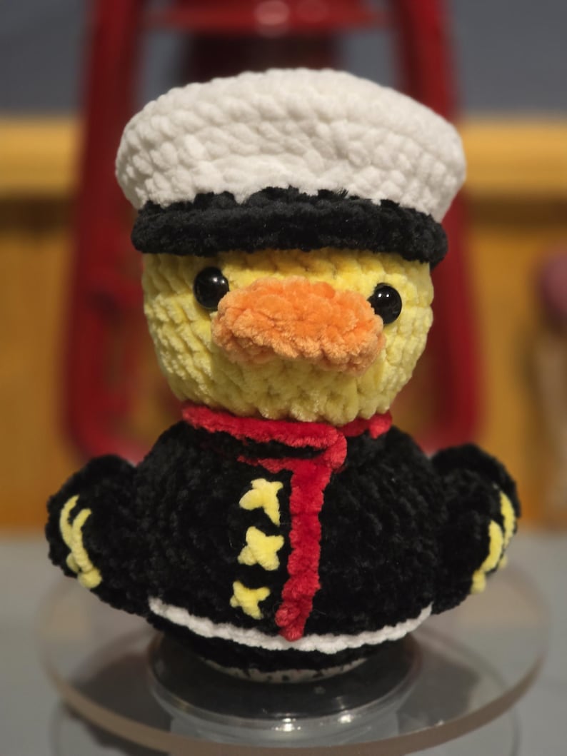 May include: A crocheted plush duck wearing a white and black sailor hat, a black jacket with yellow accents, a red scarf, and an orange beak. The duck has black plastic eyes and is crafted from soft, textured yarn in a standing, upright position.