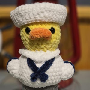 May include: A crocheted yellow duck plush toy wearing a white sailor hat and a white sailor collar with a dark blue neckerchief. The duck has black plastic eyes and an orange beak, crafted from soft, textured yarn in a small, rounded form.