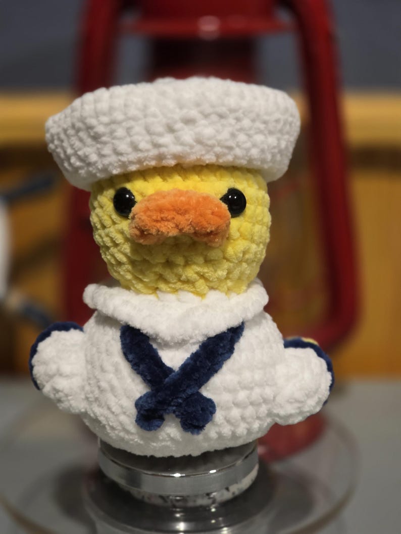 May include: A crocheted yellow duck plush toy wearing a white sailor hat and a white sailor collar with a dark blue neckerchief. The duck has black plastic eyes and an orange beak, crafted from soft, textured yarn in a small, rounded form.