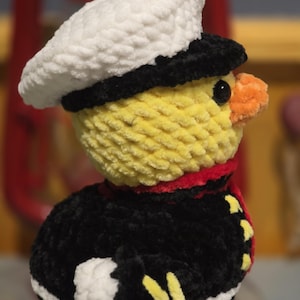 May include: A crocheted plush duckling wearing a white sailor hat with a black brim, a black uniform jacket with red trim, and yellow accents. The duckling has a bright yellow head, a small orange beak, and a black eye, crafted from soft yarn.