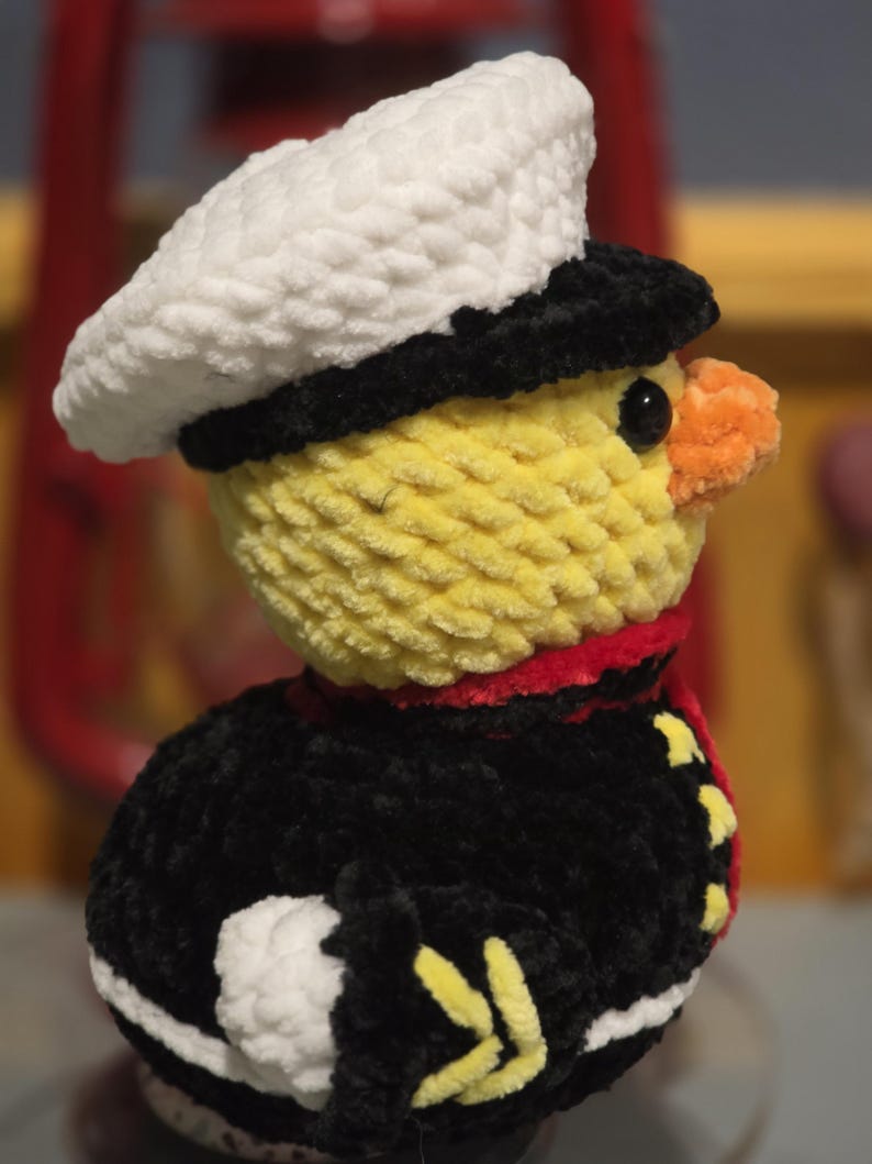 May include: A crocheted plush duckling wearing a white sailor hat with a black brim, a black uniform jacket with red trim, and yellow accents. The duckling has a bright yellow head, a small orange beak, and a black eye, crafted from soft yarn.