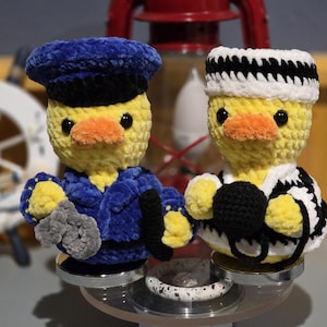 May include: Two handmade crochet duck figurines. One duck is dressed as a sailor in a blue uniform and hat, holding a gray object. The other duck is dressed in a black and white striped sailor outfit and hat, holding a black object.
