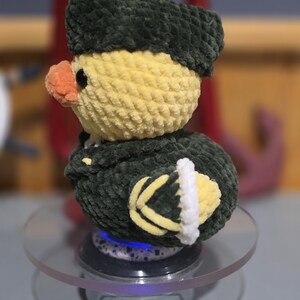 May include: A crocheted plush duck toy featuring a yellow head, an orange beak, and a dark green uniform-style jacket with a matching hat. The duck has black eyes and yellow and white detailing on the sleeves, crafted from soft yarn.