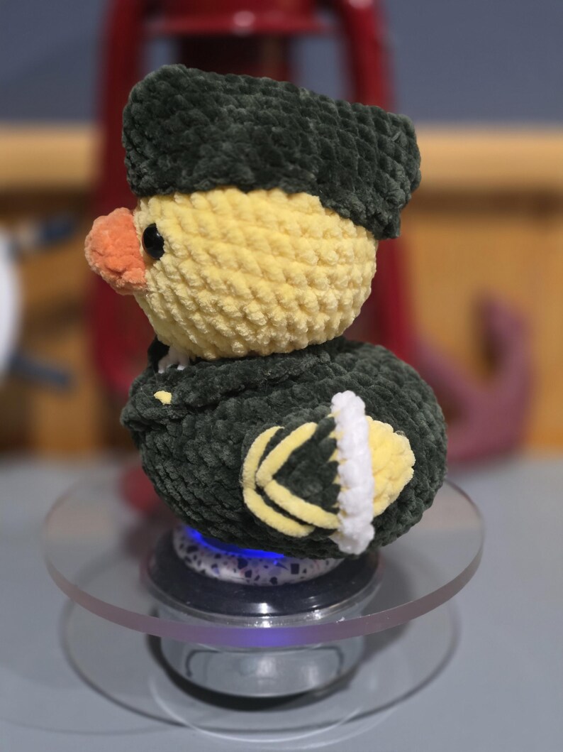 May include: A crocheted plush duck toy featuring a yellow head, an orange beak, and a dark green uniform-style jacket with a matching hat. The duck has black eyes and yellow and white detailing on the sleeves, crafted from soft yarn.