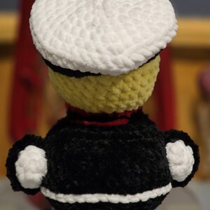 May include: A crochet doll featuring a yellow head topped with a white sailor hat, a black torso with a red collar, and white cuffs on the sleeves. The doll is crafted from soft, textured yarn and displays a distinct color-blocked design.