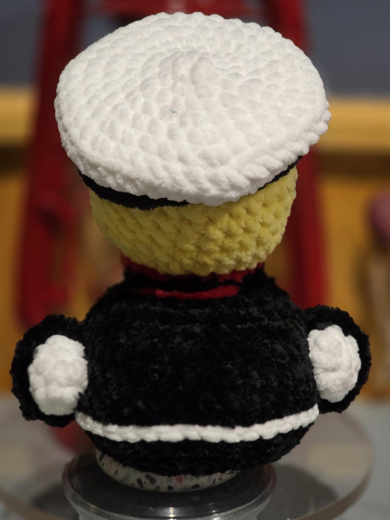 May include: A crochet doll featuring a yellow head topped with a white sailor hat, a black torso with a red collar, and white cuffs on the sleeves. The doll is crafted from soft, textured yarn and displays a distinct color-blocked design.