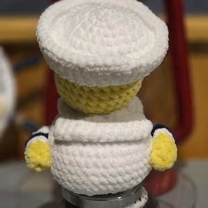 May include: A small, handmade crochet plush figure featuring a yellow head and body, wearing a white sailor-style hat and a white collared shirt with blue trim on the sleeves. The plush is crafted from soft, textured yarn with a rounded shape.