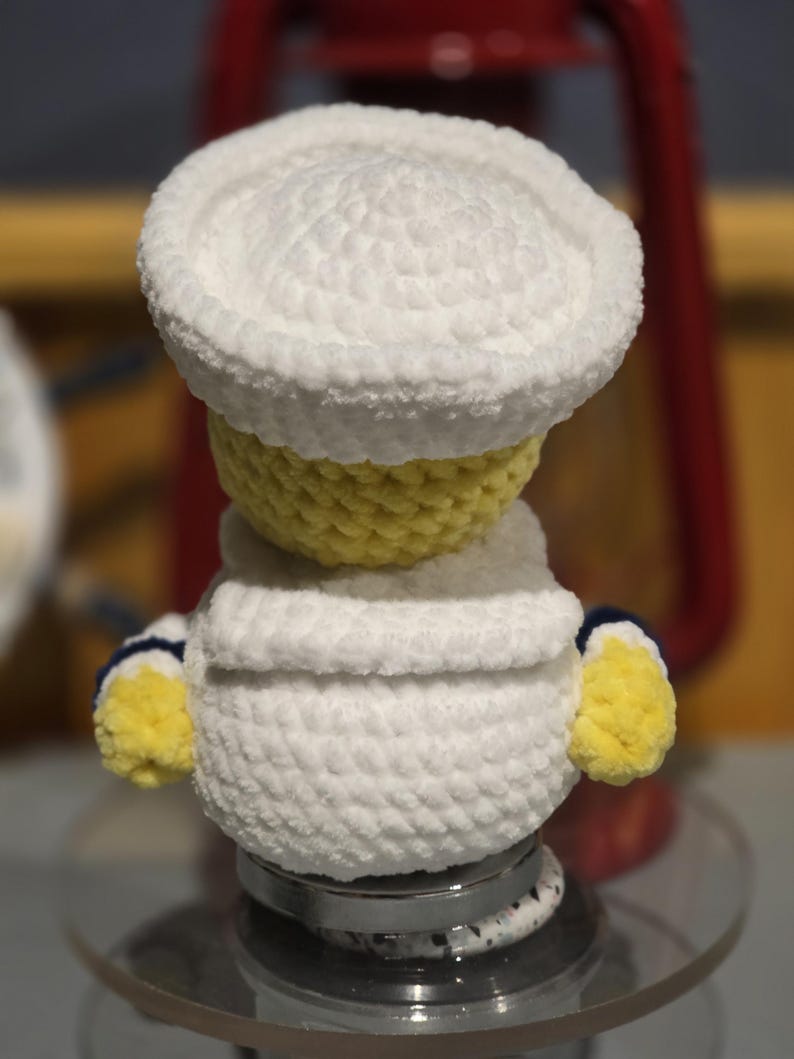 May include: A small, handmade crochet plush figure featuring a yellow head and body, wearing a white sailor-style hat and a white collared shirt with blue trim on the sleeves. The plush is crafted from soft, textured yarn with a rounded shape.