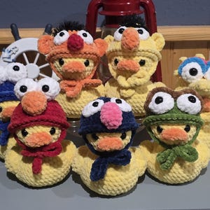 May include: A collection of handmade yellow crochet ducks, each adorned with a unique character-themed hat and scarf. The hats feature various colors, including orange, red, blue, and green, with large, expressive eyes. The ducks are arranged in a group, showcasing their whimsical designs.