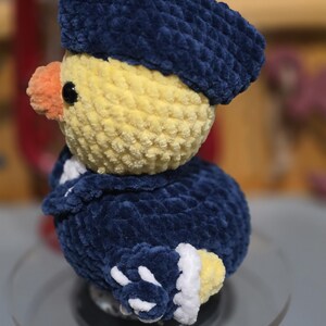 May include: A crocheted plush duckling wearing a dark blue hat and a matching dark blue jacket with white trim. The duckling has a yellow head, a small orange beak, and black bead eyes. The textured yarn creates a soft, fuzzy appearance.