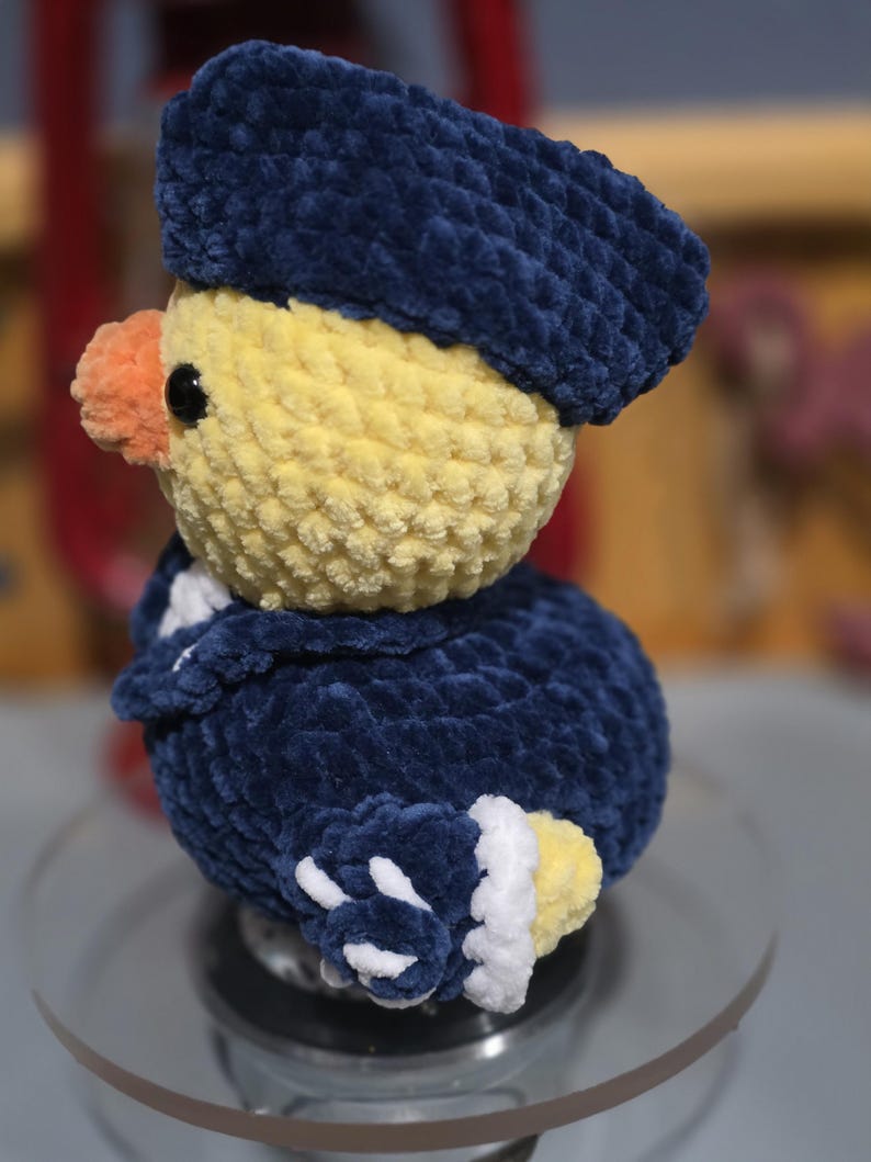 May include: A crocheted plush duckling wearing a dark blue hat and a matching dark blue jacket with white trim. The duckling has a yellow head, a small orange beak, and black bead eyes. The textured yarn creates a soft, fuzzy appearance.