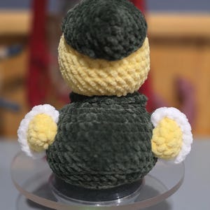 May include: A small, handmade crochet plush toy shaped like a duck. The figure features a dark green head and body, a pale yellow neck and wing accents, and white trim on the wings. The texture is soft and yarn-based with a rounded, compact form.