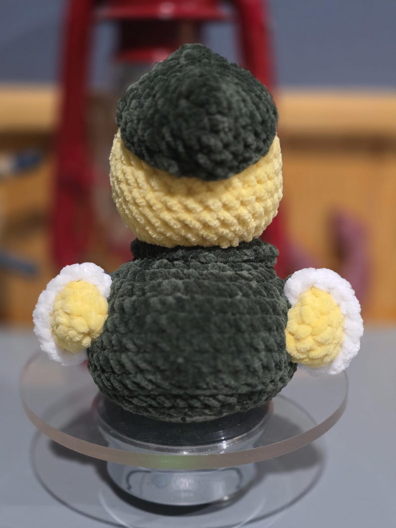 May include: A small, handmade crochet plush toy shaped like a duck. The figure features a dark green head and body, a pale yellow neck and wing accents, and white trim on the wings. The texture is soft and yarn-based with a rounded, compact form.