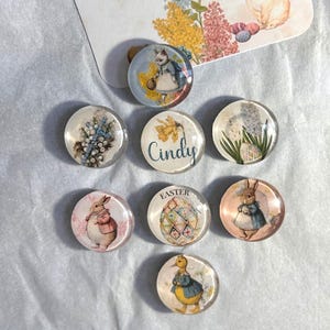 May include: A collection of round glass magnets with Easter-themed illustrations. Designs include bunnies, ducks, floral arrangements, and an Easter egg. One magnet has the name "Cindy" and another says "Easter".