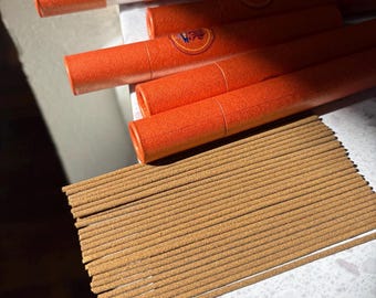 May include: An assortment of incense sticks and orange cylindrical packaging. The incense sticks are light brown and arranged in a neat row. The packaging has a textured surface and a circular emblem.
