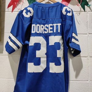 Pode incluir: Uma camisola de futebol azul com o nome "DORSETT" e o número "33" em branco. A camisola tem riscas brancas nas mangas e detalhes nos ombros. Está pendurada num cabide de madeira.