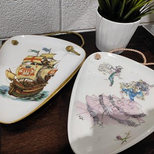 Può includere: Due piatti decorativi triangolari in ceramica. Uno presenta un'illustrazione dettagliata di una nave a vela con dettagli dorati. L'altro piatto raffigura una scena di figure con un abito rosa e un abito verde. Entrambi i piatti hanno bordi dorati e manici in corda.