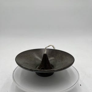 May include: A dark speckled ceramic candle holder with a conical center and a wide, shallow dish. A white wick extends from the center. The candle holder sits on a white plastic base.