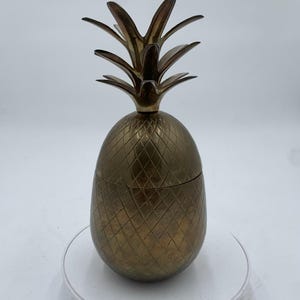 Puede incluir: Un recipiente de latón con forma de piña con tapa extraíble. El recipiente tiene una superficie texturizada con patrón de diamantes y una corona de hojas en la parte superior. La piña es un objeto decorativo.