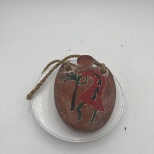 Puede incluir: Un colgante ovalado marrón con un diseño de Kokopelli en rojo, negro y verde. El colgante tiene una superficie texturizada y un cordón de yute para colgar. Descansa sobre un plato blanco.