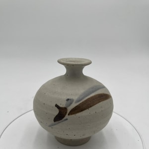 May include: A small, round, ceramic vase with a speckled, off-white finish. The vase features a short neck and a decorative design of brown and grey brushstrokes. The vase is approximately 10 cm tall.