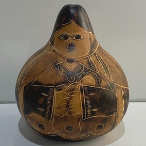 Puede incluir: Una pieza de arte de calabaza hecha a mano con un diseño tallado de una persona. La calabaza es marrón con acentos marrón oscuro, y los detalles tallados crean una apariencia texturizada. La figura está representada con un rostro y ropa detallados.