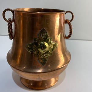 May include: A copper-colored metal vase with two looped handles and a decorative brass emblem. The handles have a twisted rope design. The vase has a wide body and a pedestal base. The brass emblem is a stylized floral design.